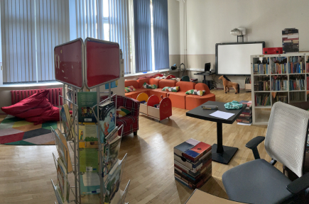 Panoramabild der Schulbibliothek. Zu erkennen sind Regale mit Büchern, ein Zeitschriftenaufsteller, Sitzsack, Couch, Smartboard.