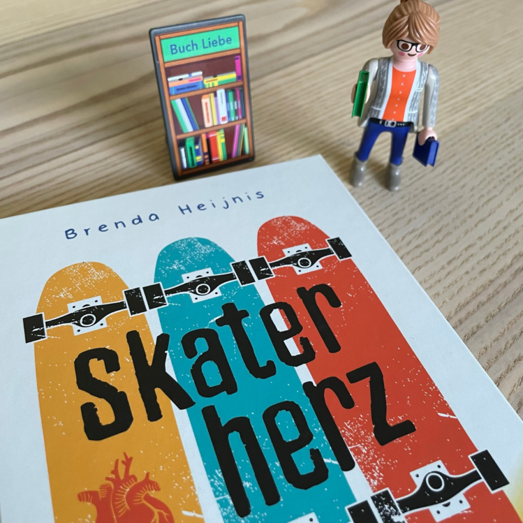 Das Cover des Buches "Skaterherz" liegt auf einem Holztisch. Das Cover ist so angeschnitten, dass nur der Titel zu sehen ist. Neben dem Buch steht eine Playmobilfigur - die Buchhändlerin.