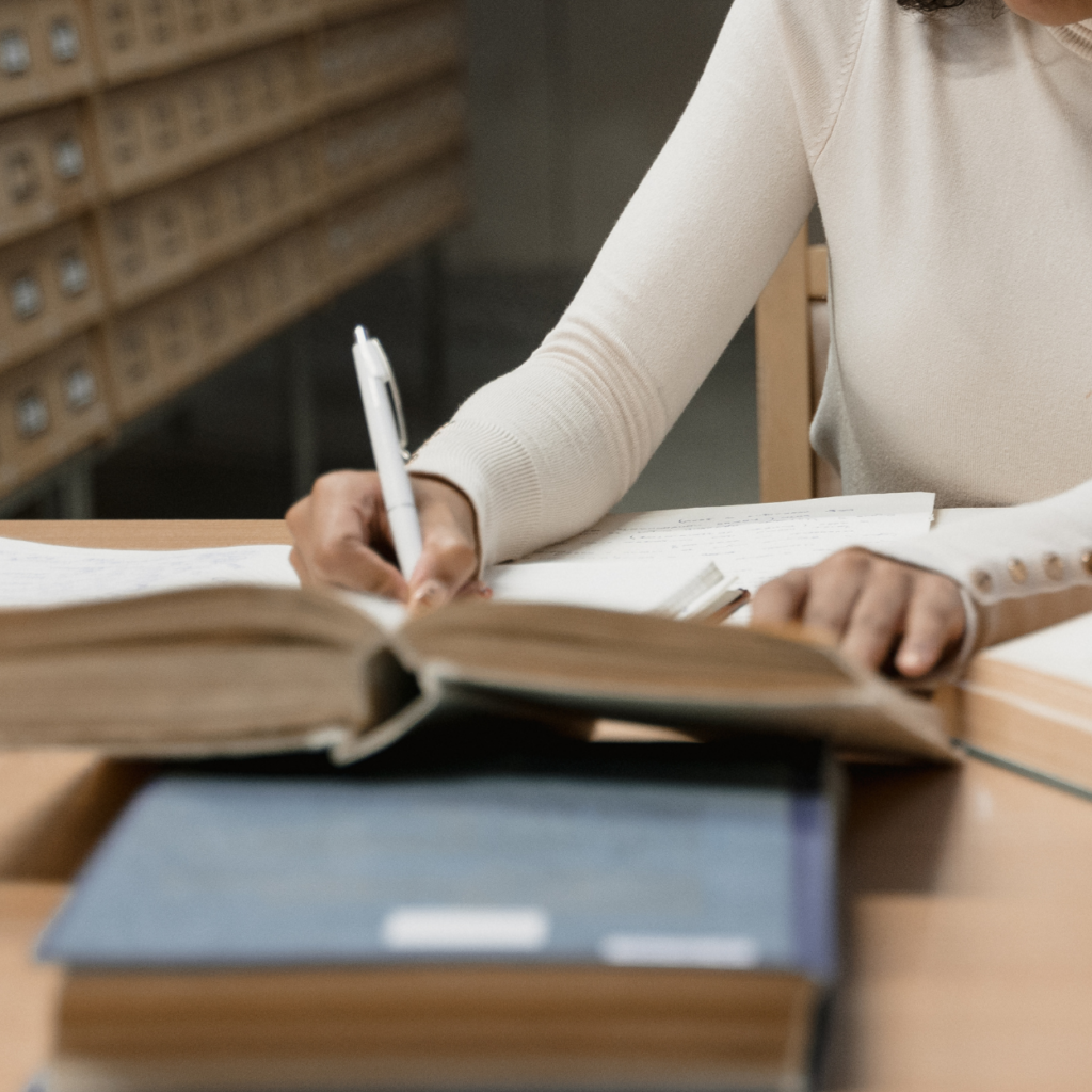 Eine Frau sitzt in einer Bibliothek. Die Frau sieht man nur angeschnitten. Im Bild sind die Hände, ein Arm und ein Teil des Oberkörpers. Auf dem Tisch vor der Frau befinden sich Bücher. Sie hat die linke Hand auf einem Buch. Mit der rechten Hand schreibt sie etwas. Im Hintergrund sieht man einen Karteischrank.