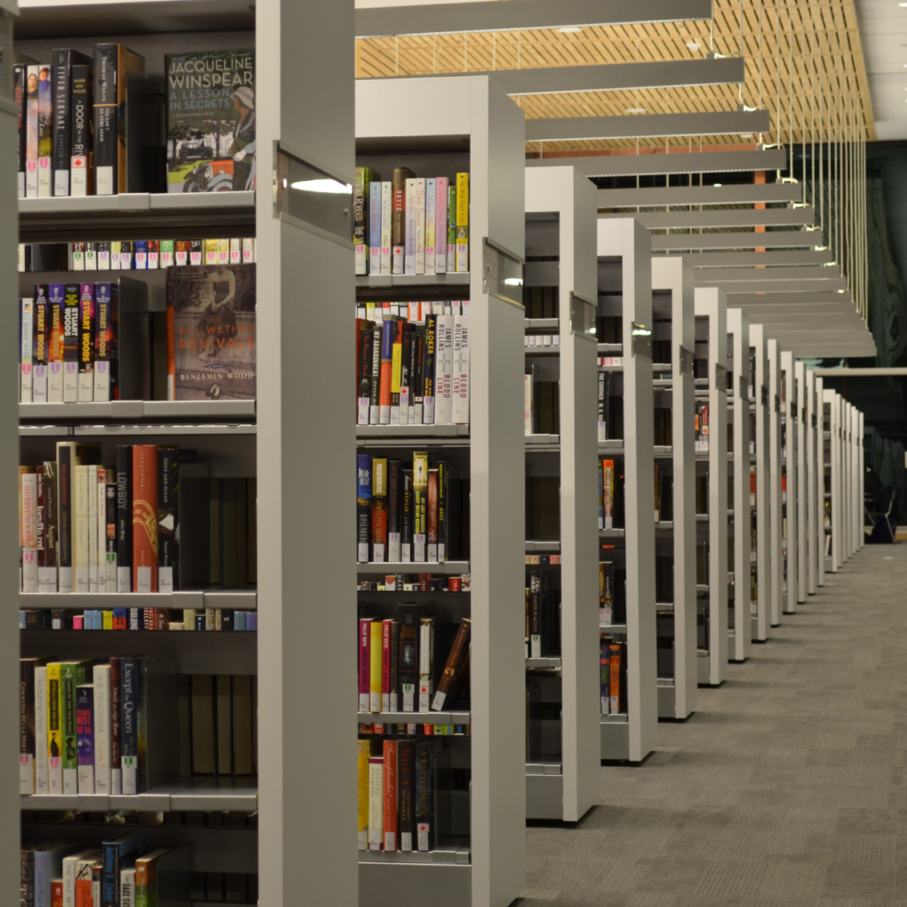 Auf dem Bild ist ein Raum in einer Bibliothek zu sehen. Es stehen viele Regale in einer Reihe. Über jedem Regal befindet sich eine Lampe.