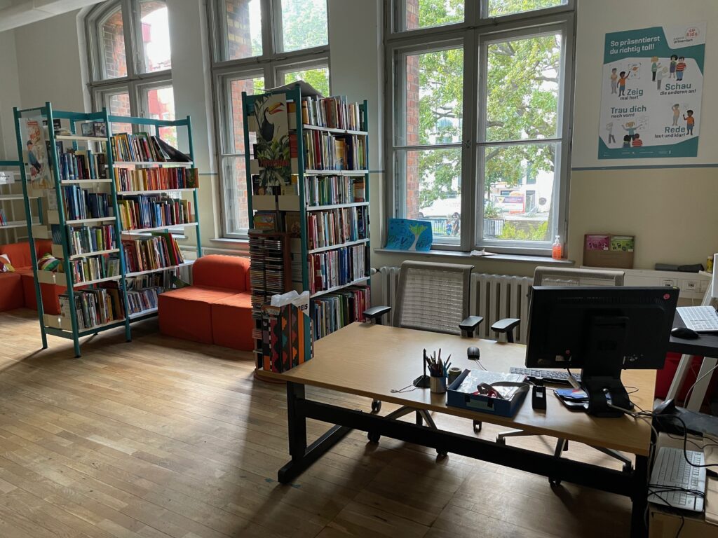 Der Blick vom Eingang der Schulbibliothek. Rechts steht der Schreibtisch mit Computer. Links sind zwei Regale, die mit Büchern gefüllt sind.