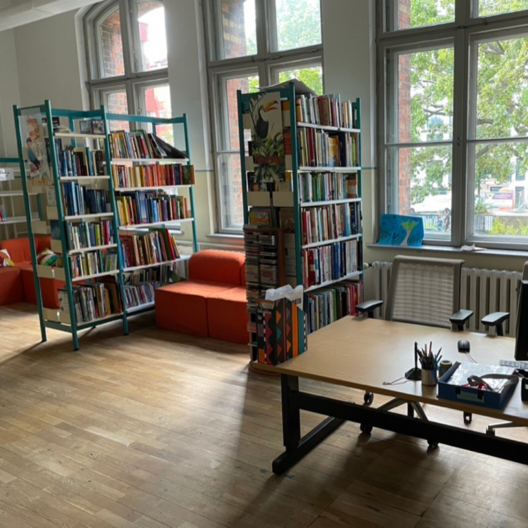 Blick in die Schulbibliothek. Rechts steht der Schreibtisch links sind zwei Bibliotheksregale. Dazwischen steht ein kleines Sofa.