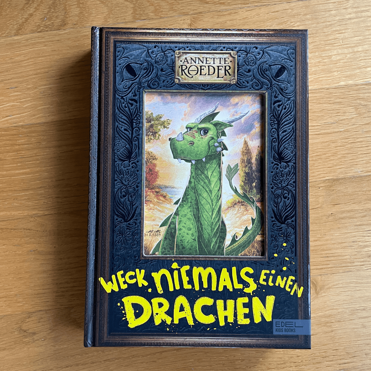 Das Buch "Weck niemals einen Drachen" liegt mit dem Cover nach oben zeigend auf einer Unterlage aus Holz. Das Buch ist schwarz, in der Mitte ist ein Drache zu sehen. Unter dem Drache steht der Titel des Buches in gelber Schrift.