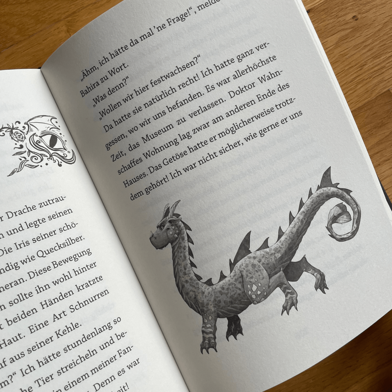 Das Buch "Weck niemals einen Drachen" liegt mit einer Innenseite nach oben zeigend auf einer Unterlage aus Holz. Auf der Buchseite ist neben dem Text ein Bild eines Drachen zu sehen.