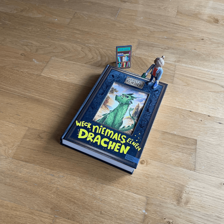 Das Buch "Weck niemals einen Drachen" liegt mit dem Cover nach oben zeigend auf einer Unterlage aus Holz. Neben dem Buch steht die Playmobilfigur "Die Buchhändlerin". Das Buch ist schwarz, in der Mitte ist ein Drache zu sehen. Unter dem Drache steht der Titel des Buches in gelber Schrift.