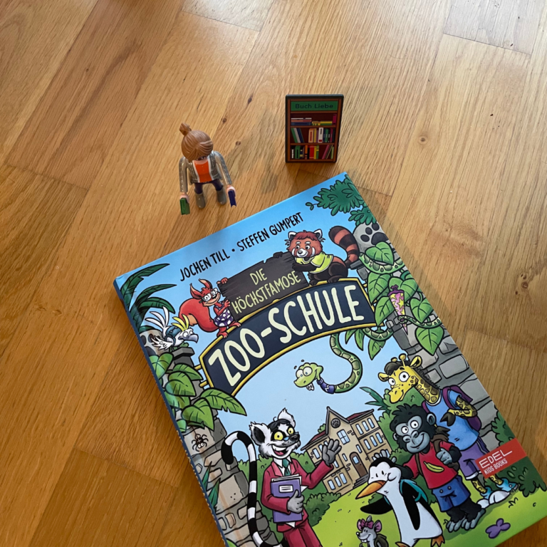 Das Cover vom Buch "Die höchstfamose Zoo-Schule" liegt auf einem Holzuntergrund. Daneben steht die Playmobilfigur "Die Buchhändlerin".