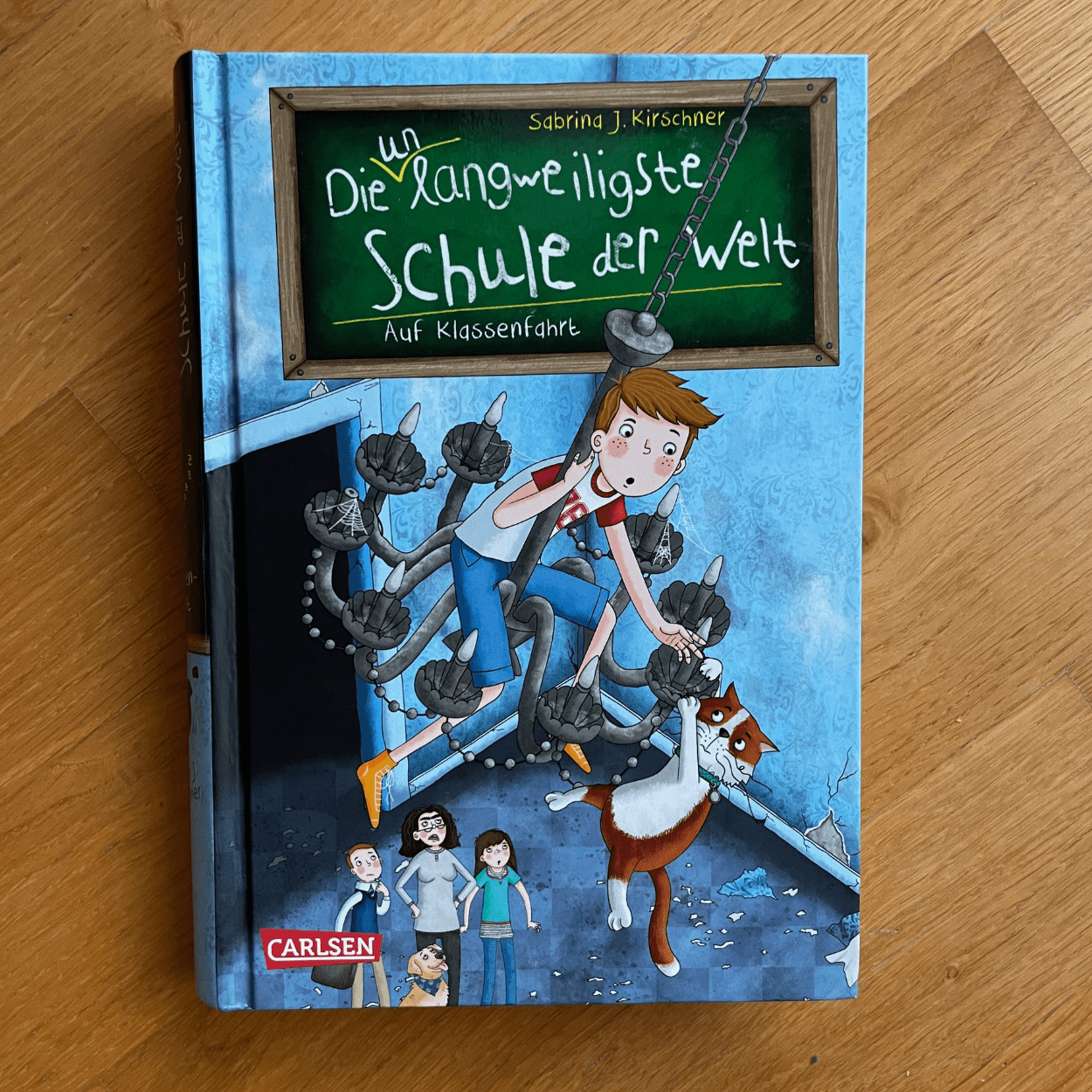 Das Buch "Die unlangweiligste Schule der Welt" Band 1 liegt mit dem Cover nach oben auf einem Holzuntergrund.