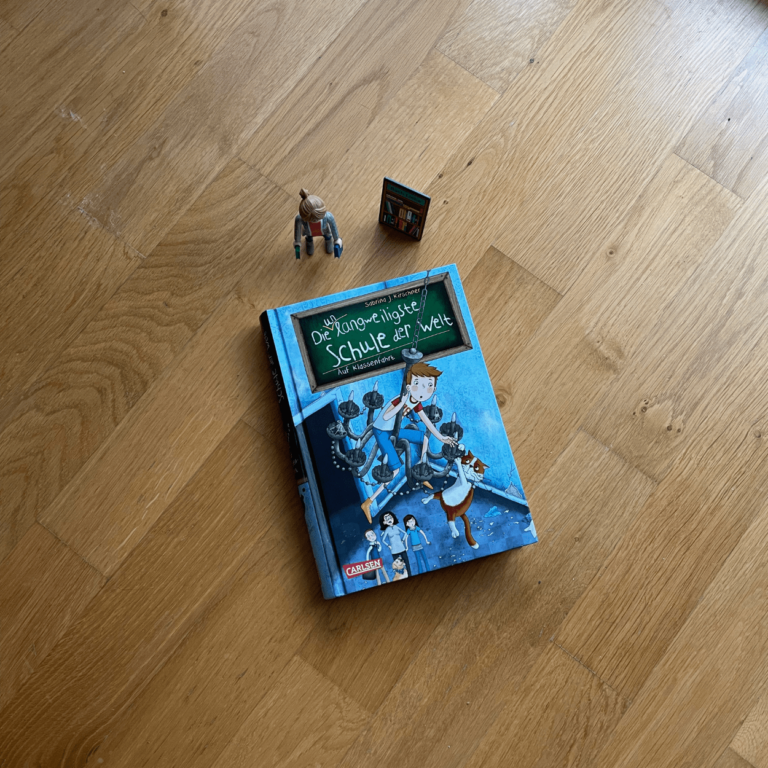 Das Buch "Die unlangweiligste Schule der Welt" Band 1 liegt mit dem Cover nach oben einem Holzuntergrund. Neben dem Buch steht die Playmobilfigur "Die Buchhändlerin".