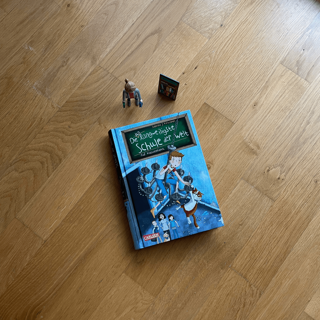 Das Buch "Die unlangweiligste Schule der Welt" Band 1 liegt mit dem Cover nach oben einem Holzuntergrund. Neben dem Buch steht die Playmobilfigur "Die Buchhändlerin".