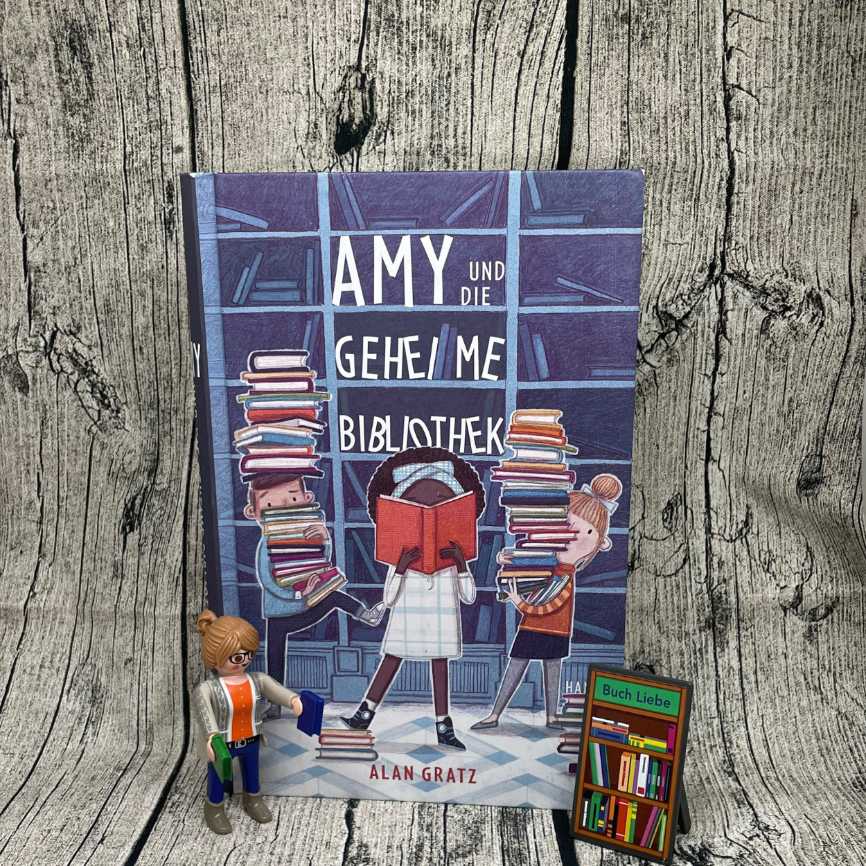 Buch "Amy und die Geheime Bibliothek" steht vor Hintergrund in Holzoptik.