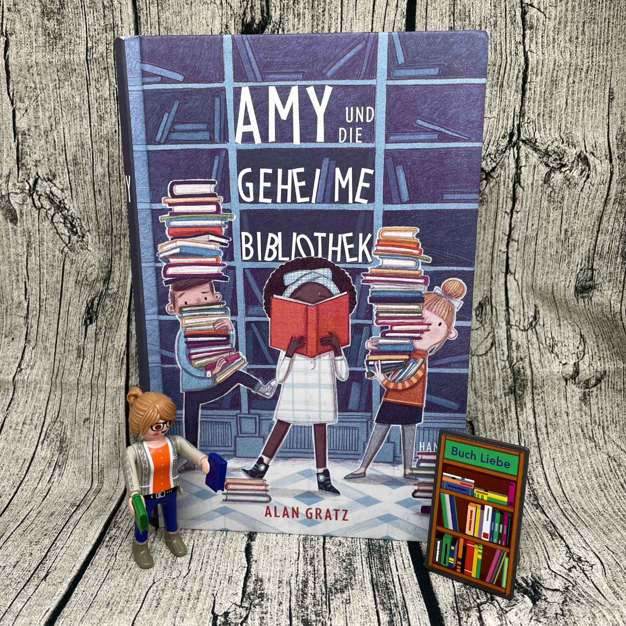 Buch "Amy und die Geheime Bibliothek" steht vor Hintergrund in Holzoptik. Links vor dem Buch steht die Playmobilfigur Die Buchhändlerin. Rechts vor dem Buch steht das zur Figur gehörende Buchregal.